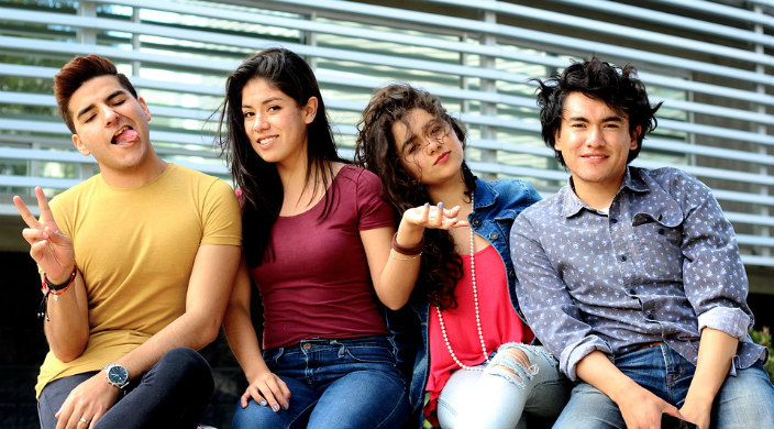Four college aged friends in colorful clothing sitting on a bench together smiling and making funny faces